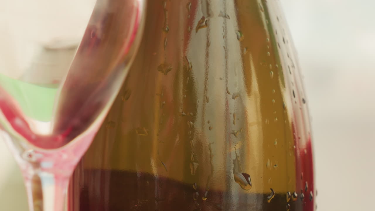 close up botol dan gelas anggur basah, tetesan embun menempel pada botol dingin sementara tepi anggur merah muda dan batang ramping menciptakan suasana romantis yang intim, lampu latar lembut dan fokus dangkal menghasilkan kehangatan