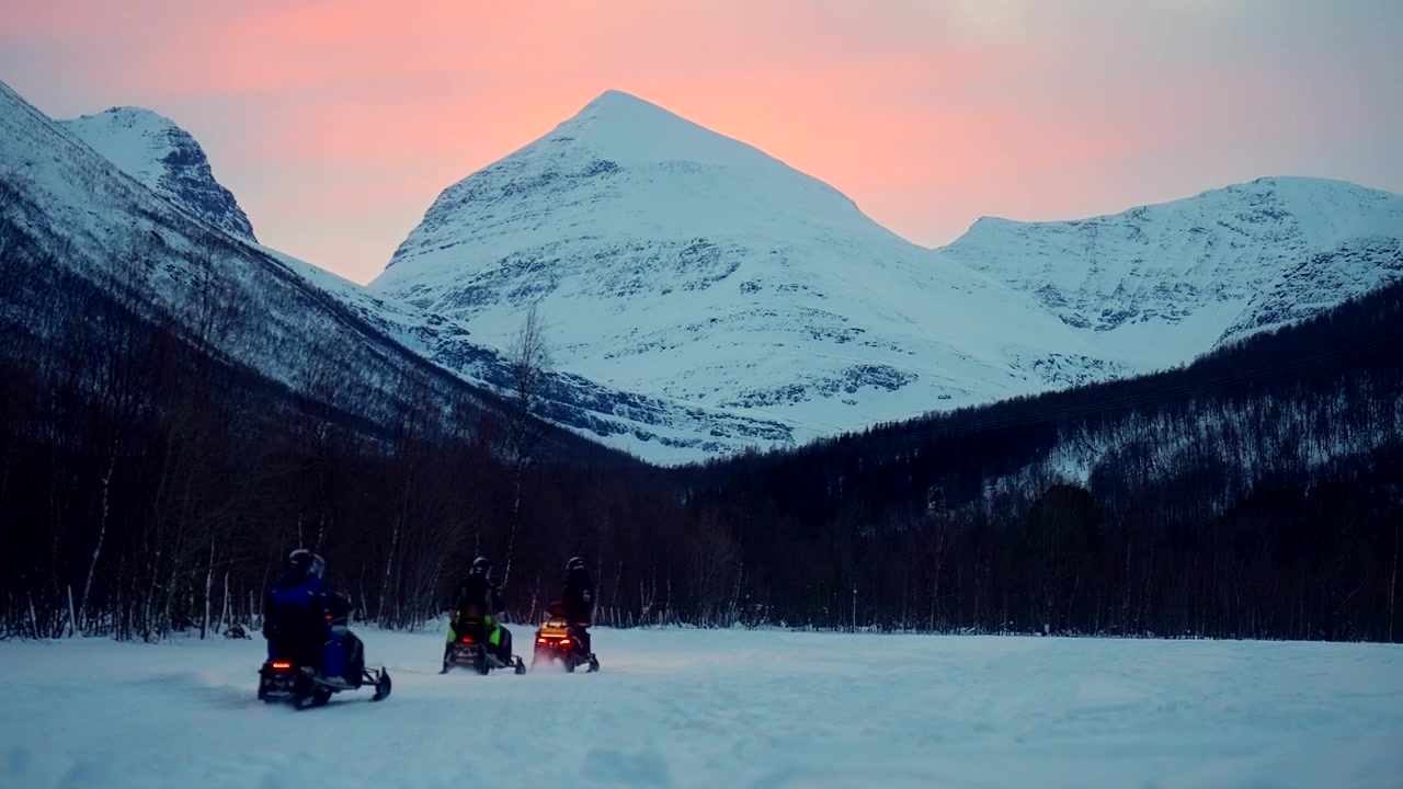 Motoslitte in escursione attraverso terreno innevato e montagne sotto un cielo notturno polare.