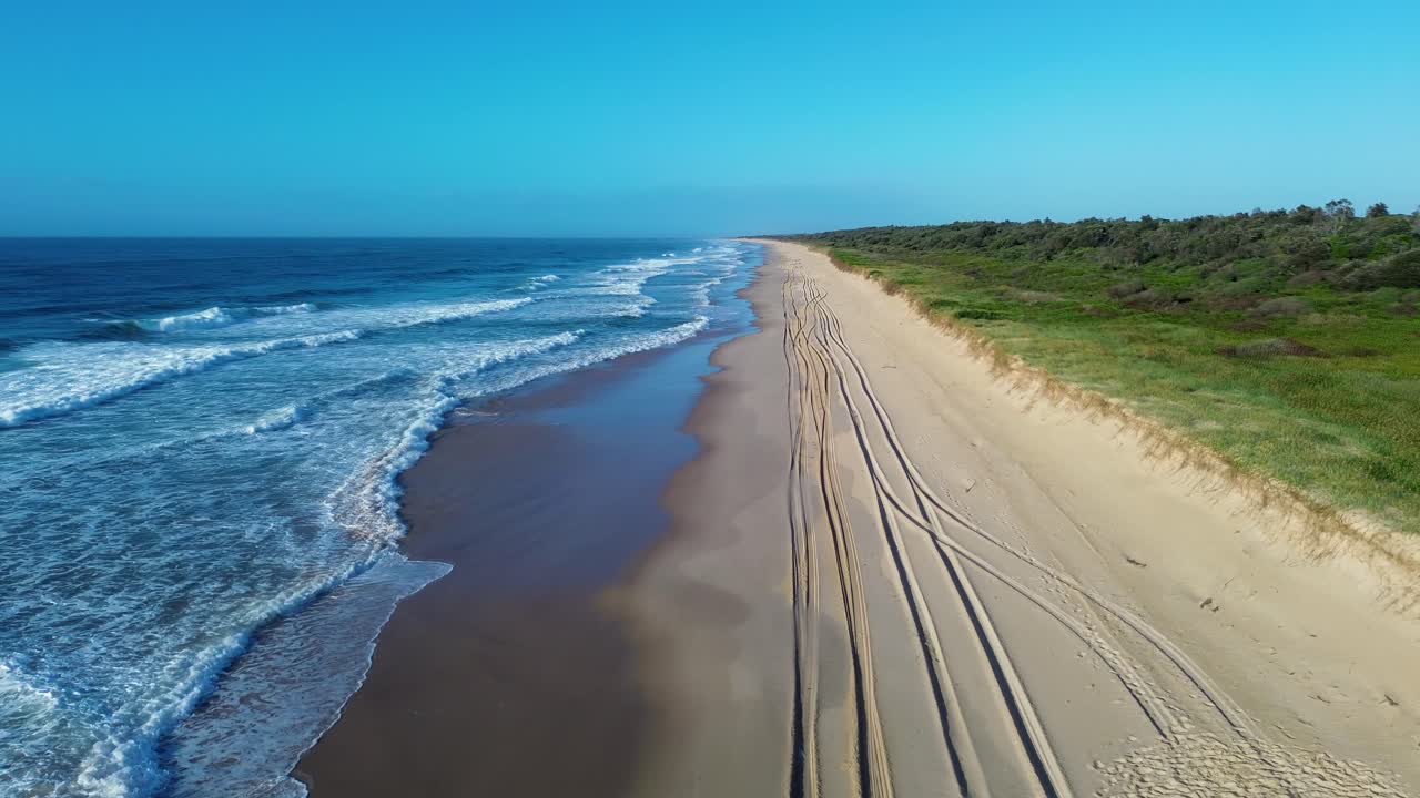 Avustralya, NSW Ulusal Parkı Harrington’da kum tepeleri boyunca araba lastiği izleri olan ve yakındaki çalılık orman ağaçlarının bulunduğu, okyanus dalgalarının kumsalda kıyı şeridini kırdığı drone hava manzarası