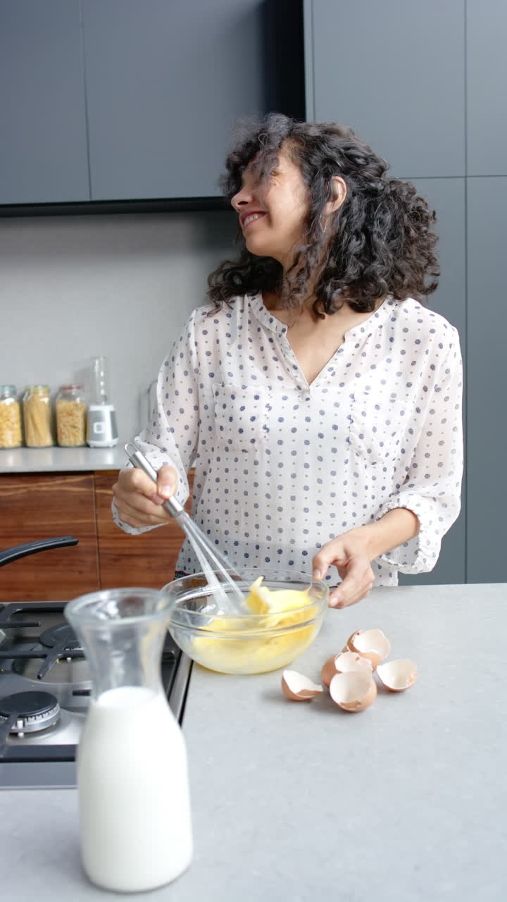 Vertical video of happy mature caucasian woman whisking eggs in kitchen at home, slow motion