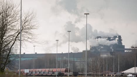 industrial plant with smokestacks emitting steam against a cloudy sky, train cargo in the foreground