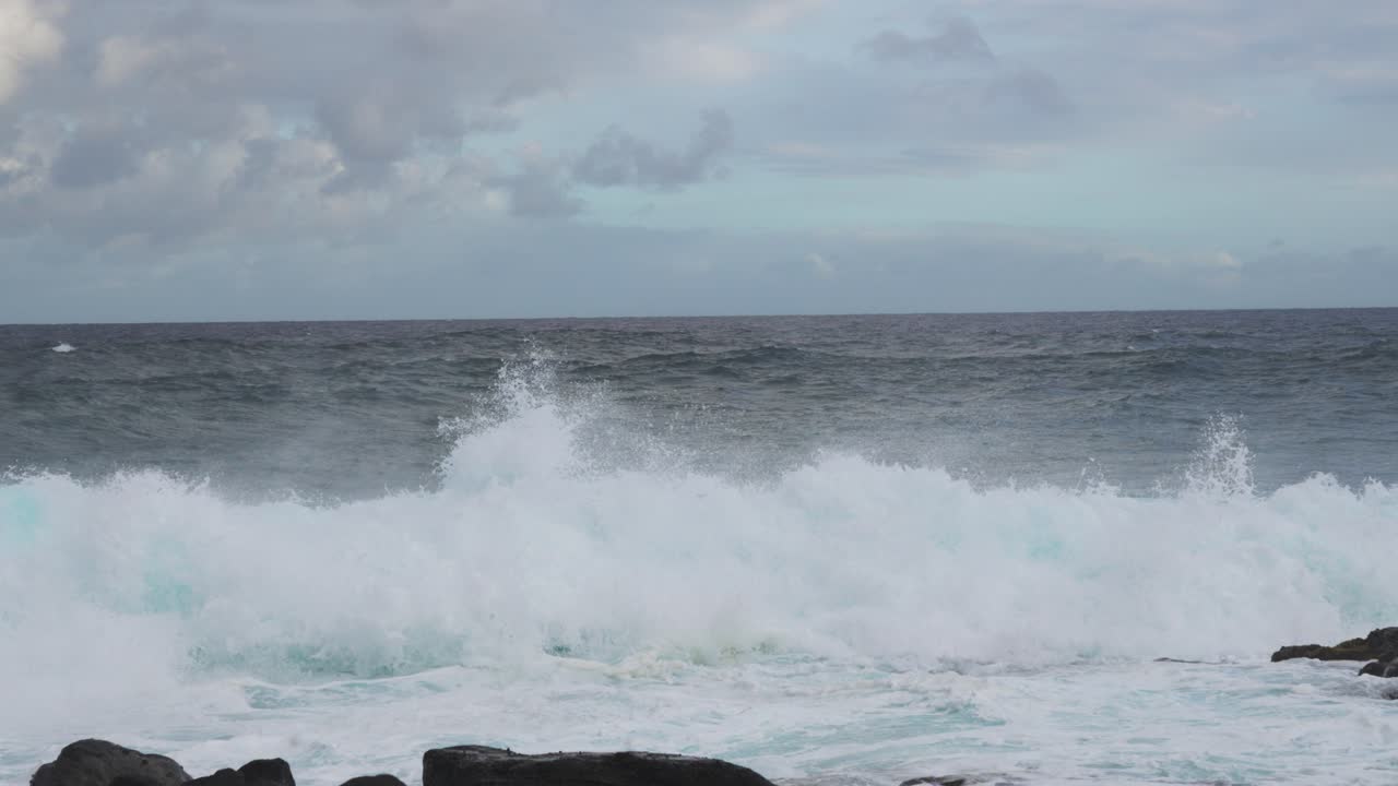 A backlit turquoise wall of swell collapses near shore, erupting into boiling whitewater around dark rocks, as clouds drift overhead and steady onshore wind heaves the horizon