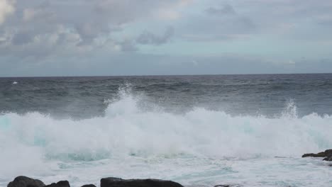 a backlit turquoise wall of swell collapses near shore, erupting into boiling whitewater around dark rocks, as clouds drift overhead and steady onshore wind heaves the horizon