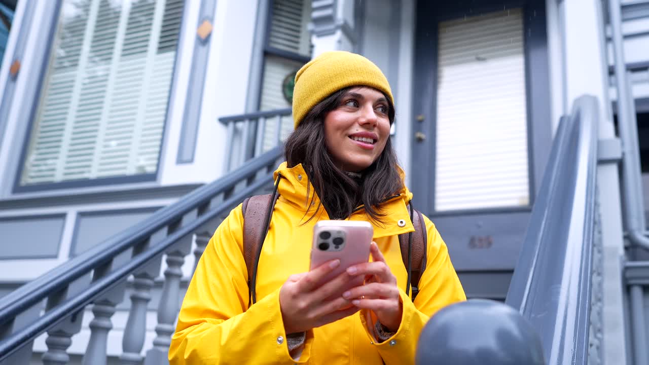 Blije jonge vrouw in gele regenjas gebruikt smartphone op stadstrappen