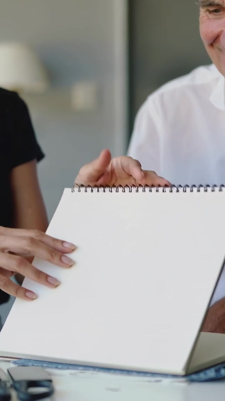 Vídeo vertical: Apontando para uma página em branco, mulher à esquerda com blusa escura virando o caderno de esboços para revisão na mesa.
