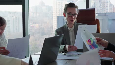 Active-meeting-of-businesswomen-at-a-table-in-the-office-to-solve-case-and-task.-A-confident-middle-aged-girl-in-glasses-and-a-business-suit,-the-leader-of-a-businesswoman-group-sits-at-the-table-and-takes-work-and-plans-schedules