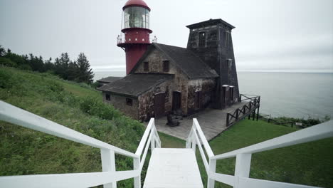 tilting up from rustic white country style stairs to reveal a red lighthouse and rustic country barn style building on the coast during an overcast day