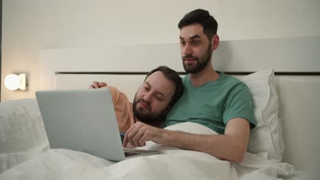 Male-gay-couple-spend-time-at-home-lying-in-bed,-using-laptop-computer