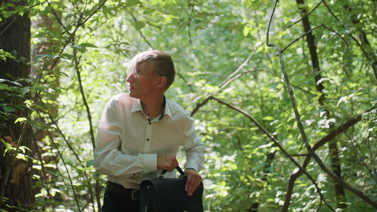 Close view of young man in white shirt carrying backpack while bending carefully and walking through dense green forest under daylight, moving cautiously between branches and trees