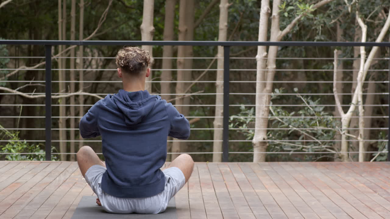 Meditoi ulkona, hupparipäinen mies istuu risti-istunnossa puisella terassilla.