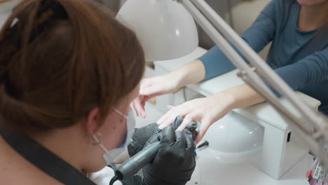 overhead view of nail technician with tied blond hair wearing face mask and apron, carefully filing client nails with electric file under desk lamp in modern salon environment
