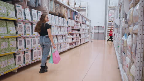 female shopper walking through supermarket aisle, looking at bedding products on shelves while other shoppers pass by in background, store shelves stocked with duvet covers and bedspreads