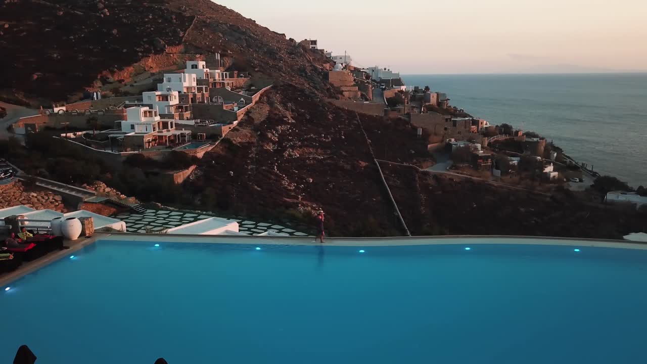 Aerial View of Woman in Summer Dress Walking on Border of Infinity Swimming Pool in Exclusive Greek Island Resort on Sunset