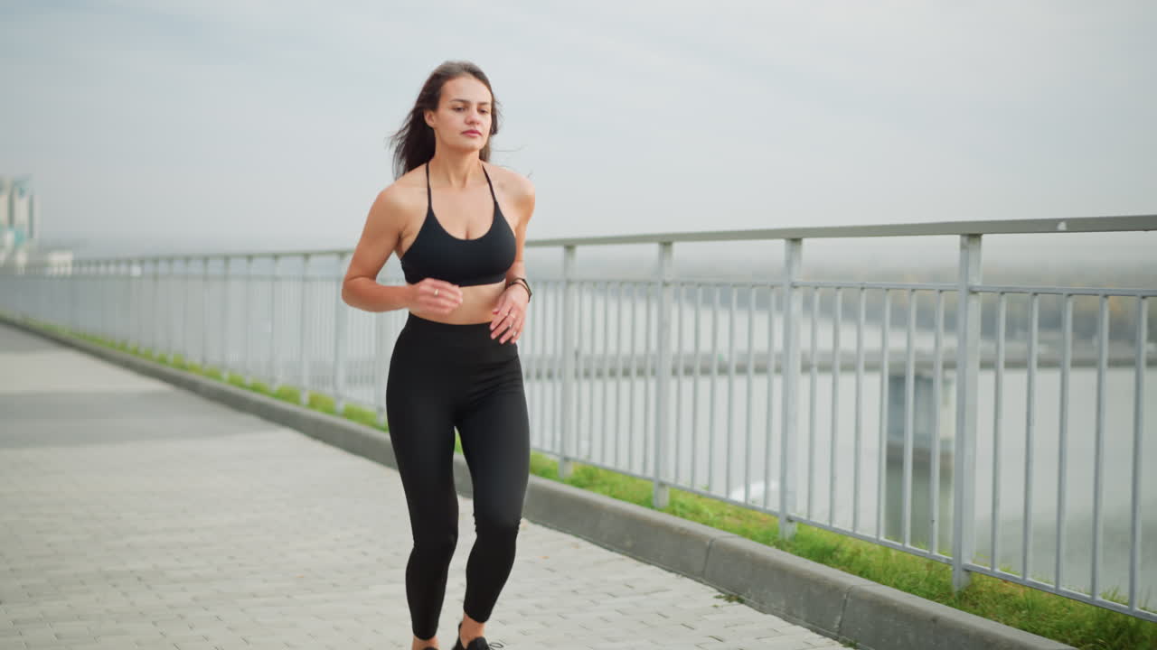 Giovane ragazza in top nero a collo alto e leggings neri che corre accanto a una recinzione di ferro con uno sfondo panoramico che presenta un ponte e un fiume, mostrando uno stile di vita sano e attivo in un contesto urbano