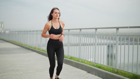 young girl in black neck top and black leggings running beside an iron fence with a scenic background featuring a bridge and river, showing a healthy, active lifestyle in an urban setting