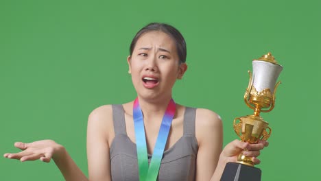 close up of wondering asian woman with a gold medal looking at trophy doubtfully on green screen background in the studio