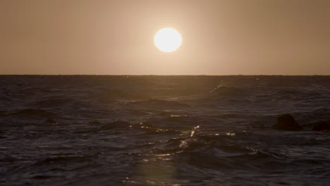 shot from the beach in hawaii, looking out onto the ocean, as waves crash in front of the setting sun