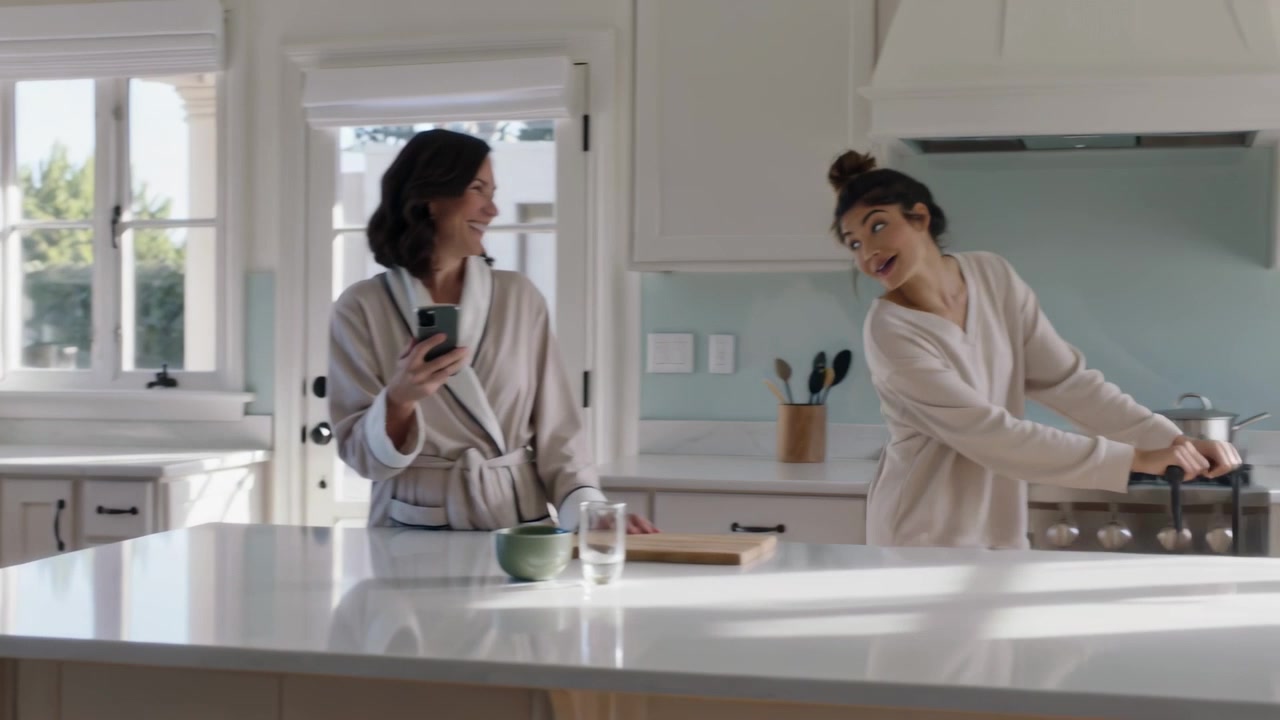 Donna sorridente che tiene uno smartphone, mentre un'amica in vestaglia trascina una valigia vedendo il telefono nella cucina di casa.