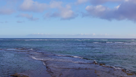 drone-shot-quickly-arises-from-the-sandy-beach-to-see-the-vast-blue-hue-of-the-endless-Pacific-ocean-and-the-blue-sky-with-puffy-clouds-at-Diamond-Head-Oahu,-Hawaii