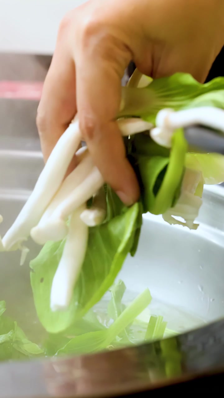 Close-up of hand dropping leafy greens and mushrooms into steaming tabletop hotpot broth