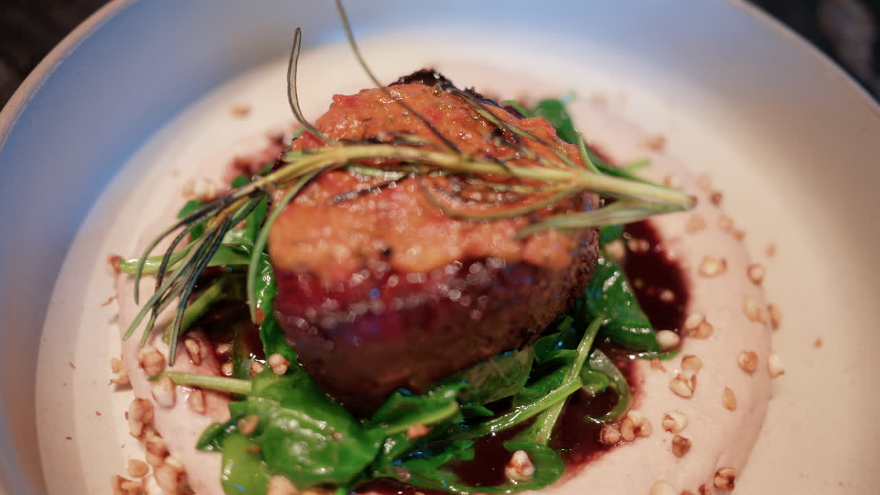 Close up of a gourmet meat dish served with sauce, greens, and rosemary garnish on a white plate