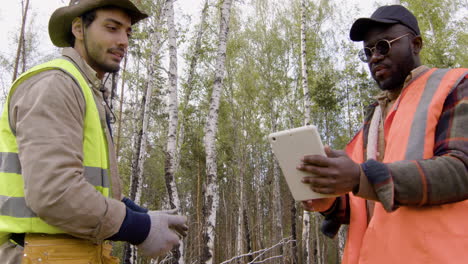 Vista-Inferior-De-Un-Activista-Caucásico-Y-Un-Hombre-Afroamericano-Viendo-Algo-En-Una-Tableta-En-El-Bosque-Mientras-Deciden-Dónde-Plantar-árboles