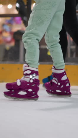 vista cercana de dos patinadores en una pista de hielo, uno en patines púrpuras con pantalones verdes menta, el otro en patines azules con pantalóns negros, el fondo muestra una vista borrosa de la gente