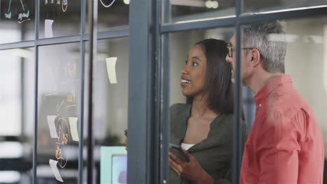 happy diverse male and female colleague brainstorming, writing on glass wall in office, slow motion