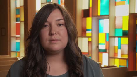 Close-up-of-a-pretty-young-ethnic-female-staring-sadly-at-the-camera-inside-a-church-sanctuary-while-sitting-in-a-pew