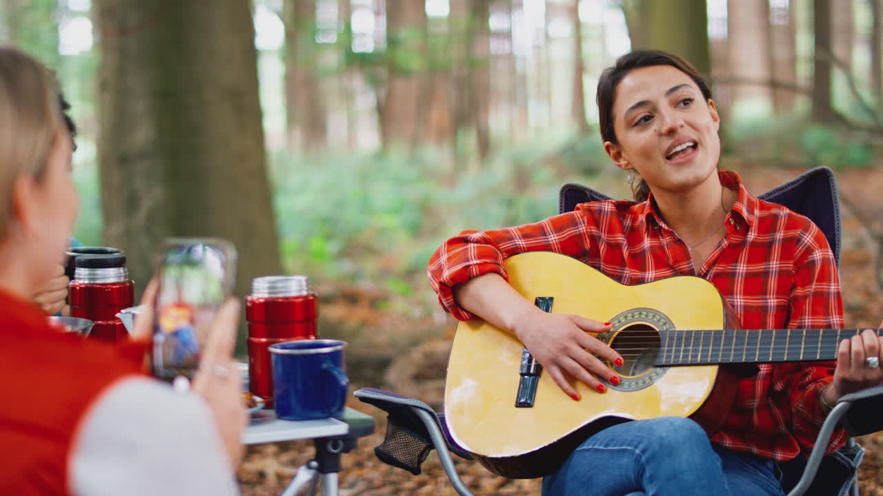 mujeres en vacaciones de campamento en el bosque comiendo comida y filmando amigo cantando junto a la guitarra