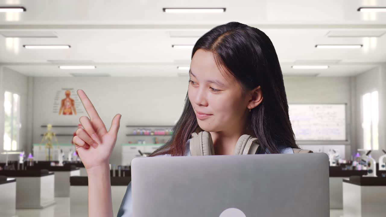 백을 들고 있는 아시아 십대 소녀 학생이 과학 실험실에 서 있는 동안 노트북을 사용하고 을 가리키고 있습니다.