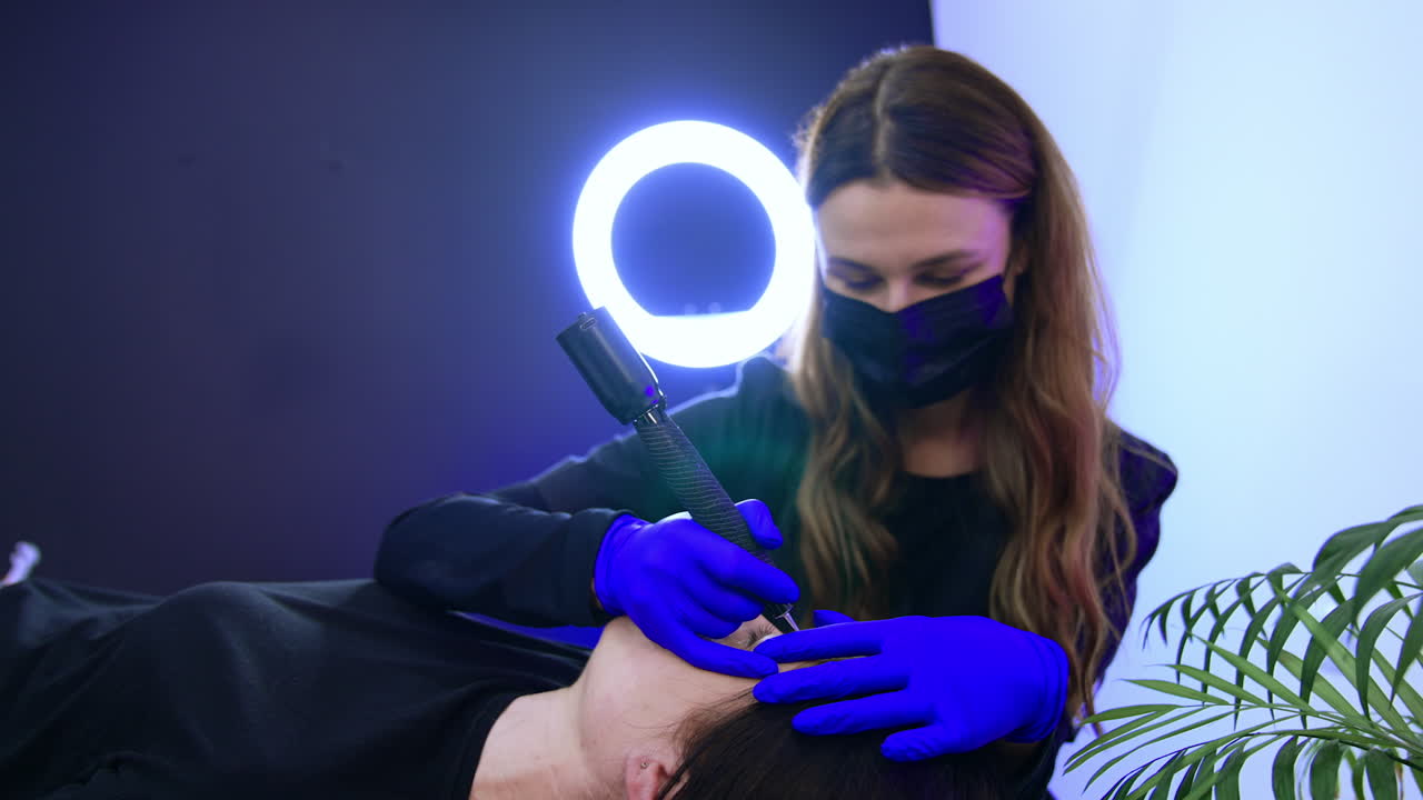 Long-haired brunette brow master in black mask applies special tool for eyebrow make up. Permanent make up procedure for a ladies in salon.