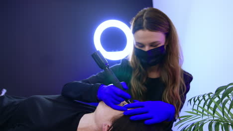 long-haired brunette brow master in black mask applies special tool for eyebrow make up. permanent make up procedure for a ladies in salon.