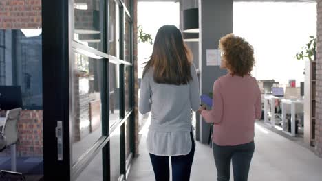 rear view of biracial female colleagues walking through office talking, slow motion