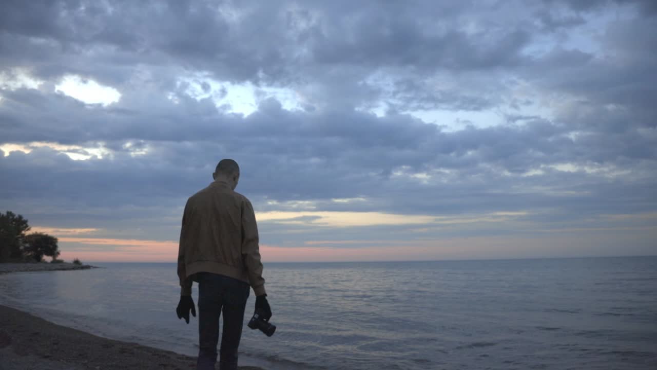 Caucasian Man Walking Towards The Edge Of The Shore With His Camera - medium shot