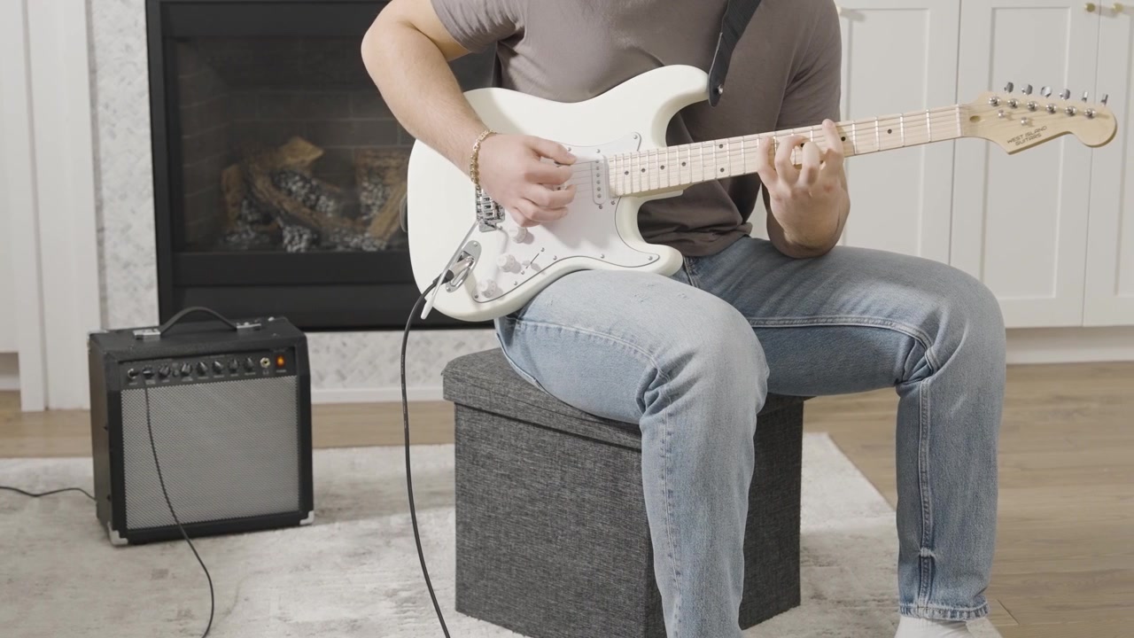 Genç bir adamın oturma odasında, yanında küçük bir amfi ile beyaz bir elektro gitar çalarken çekilen kaydırma görüntüsü.