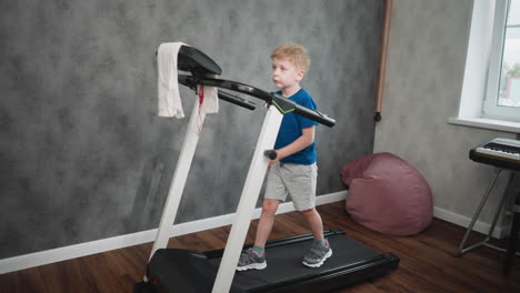 boy on treadmill gentle walking for workout in cozy room with bean bag and keyboard near window grey textured wall wooden floor towel hung on console showing casual child exercise routine