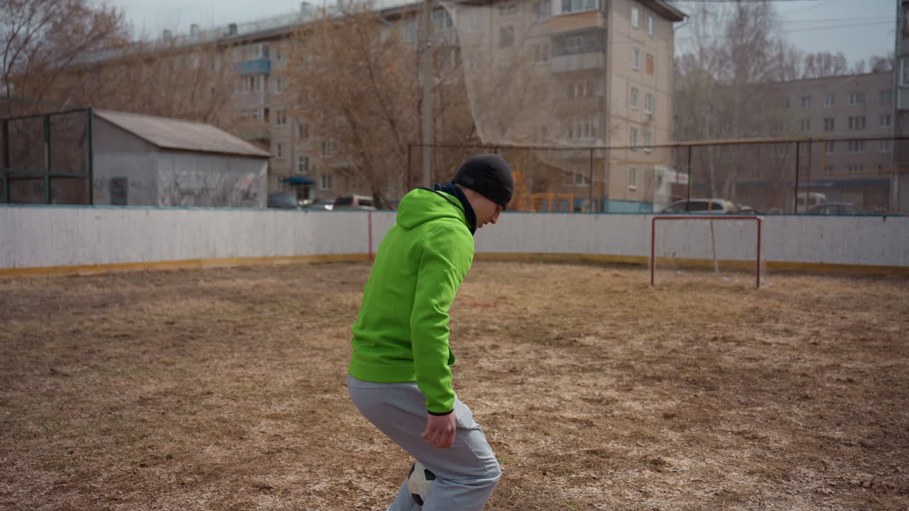 Atleta controla el balón con destreza; persona con sudadera luminosa maneja el balón con habilidad en el césped; individuo vestido con una sudadera de color neón practica movimientos precisos cerca de una portería rústica