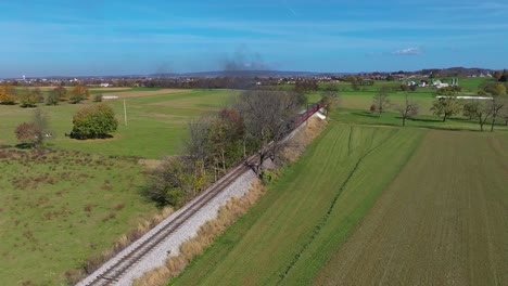 an aerial view of a steam train traveling over a small bridge as it travels thru country farmlands on a sunny fall day