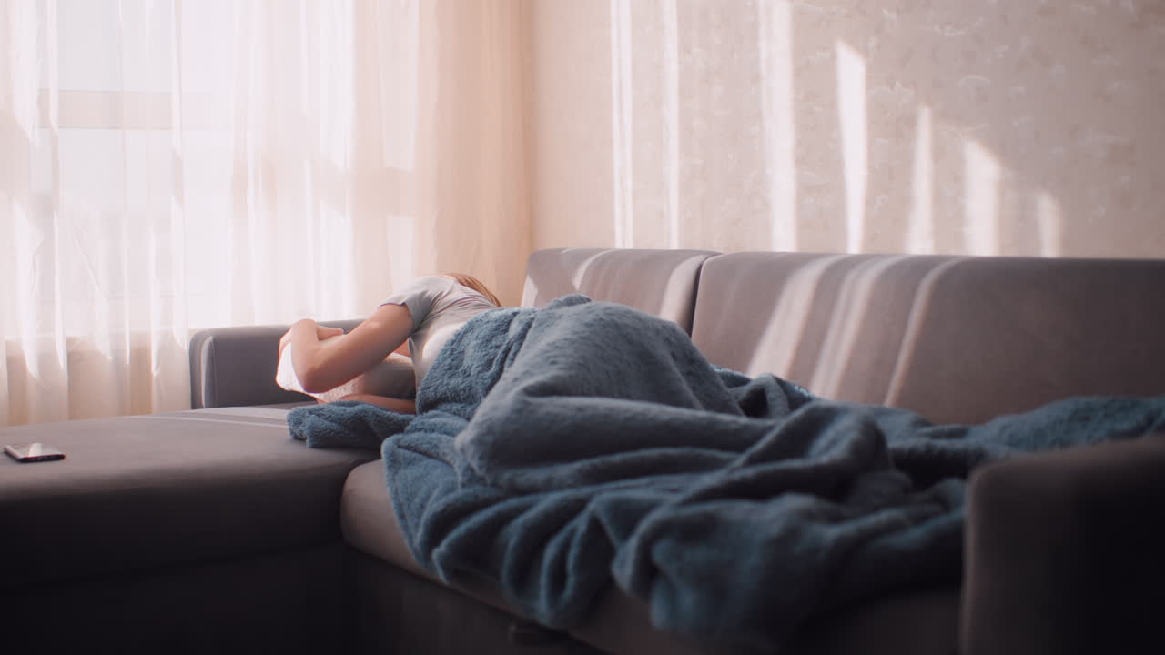 Exhausted woman curled up on couch after long workday, using pillow to cover face from morning light, soft blue blanket wrapped around her as mobile phone rests nearby in sunlit quiet room