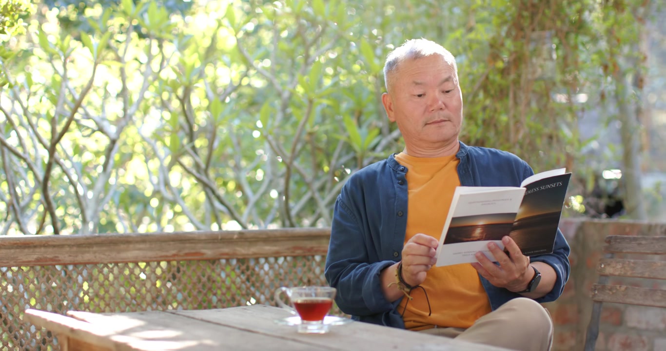 ポーチでくつろぎながらお茶を飲み、雑誌のページをめくる高齢のアジア人男性、コピースペース。