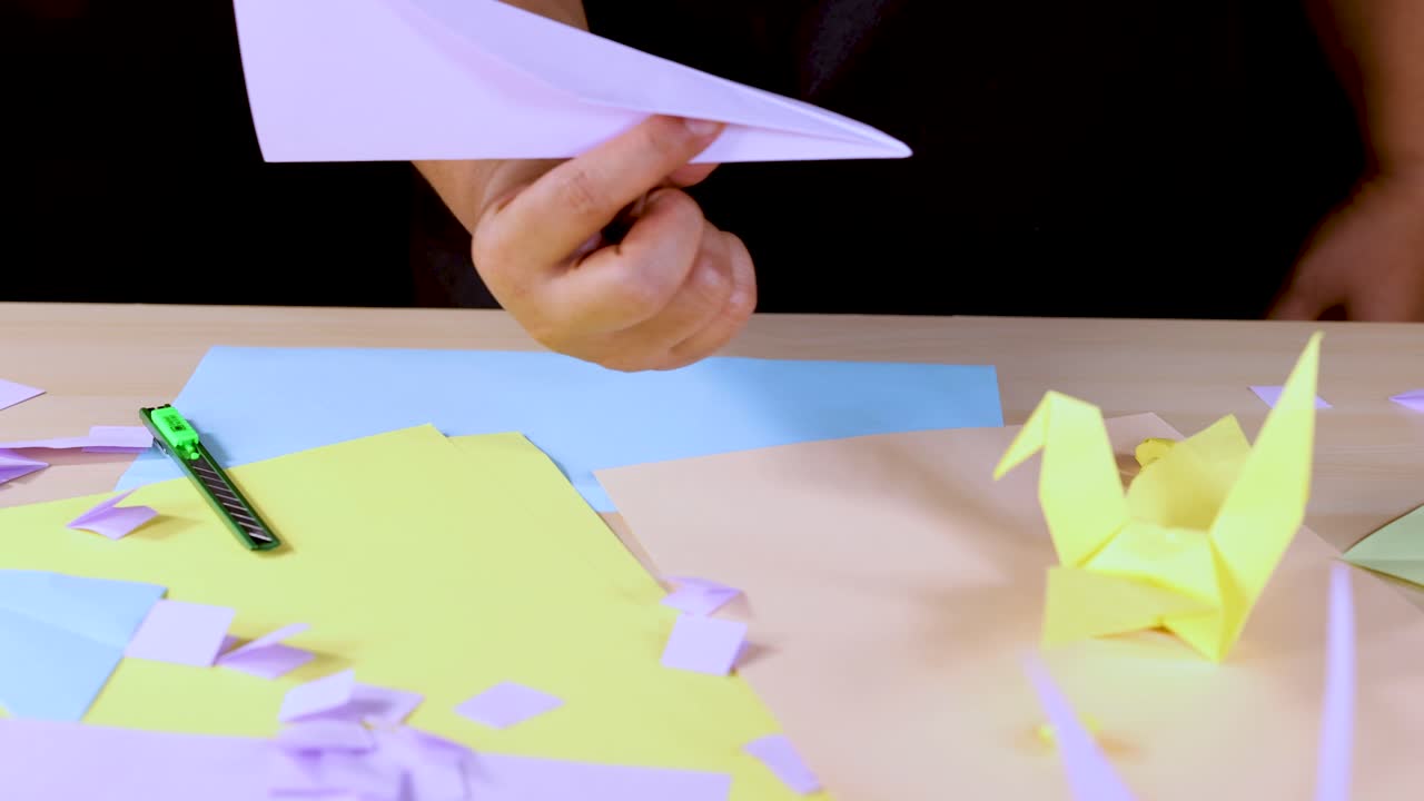 Person displays paper airplane above colorful origami workspace with bright, even lighting and static camera