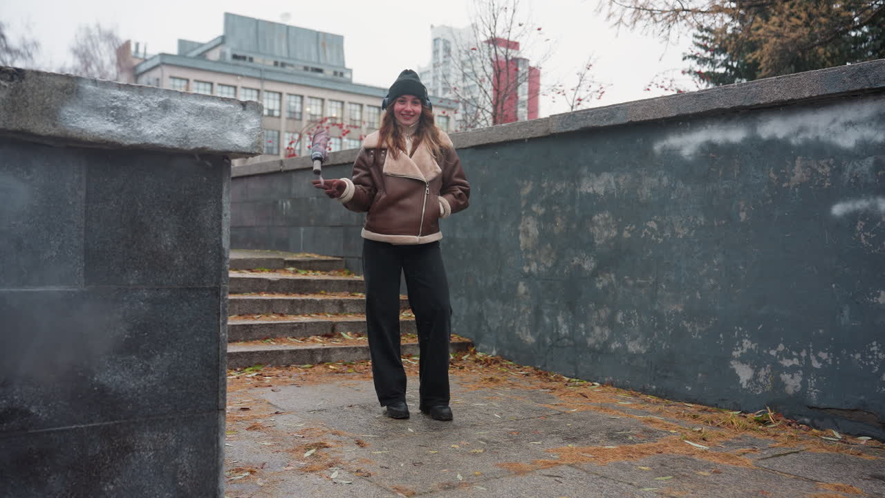 Cheerful lady wearing black knit cap, brown shearling jacket, black trousers, standing outdoors with one hand in pocket while swinging umbrella playfully on concrete path with autumn leaves scattered