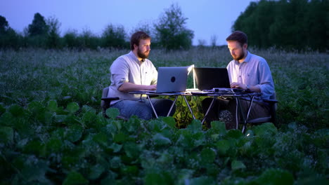 Two men working on laptops outdoors in a field at night