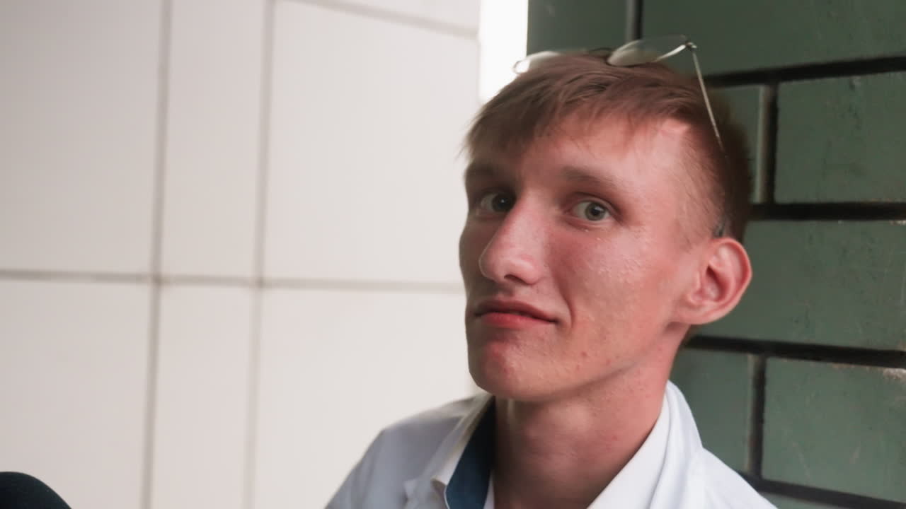 Close view of young man wearing white shirt with glasses resting on head, sitting outdoors near wall, looking directly at camera with calm and thoughtful expression, capturing natural human detail