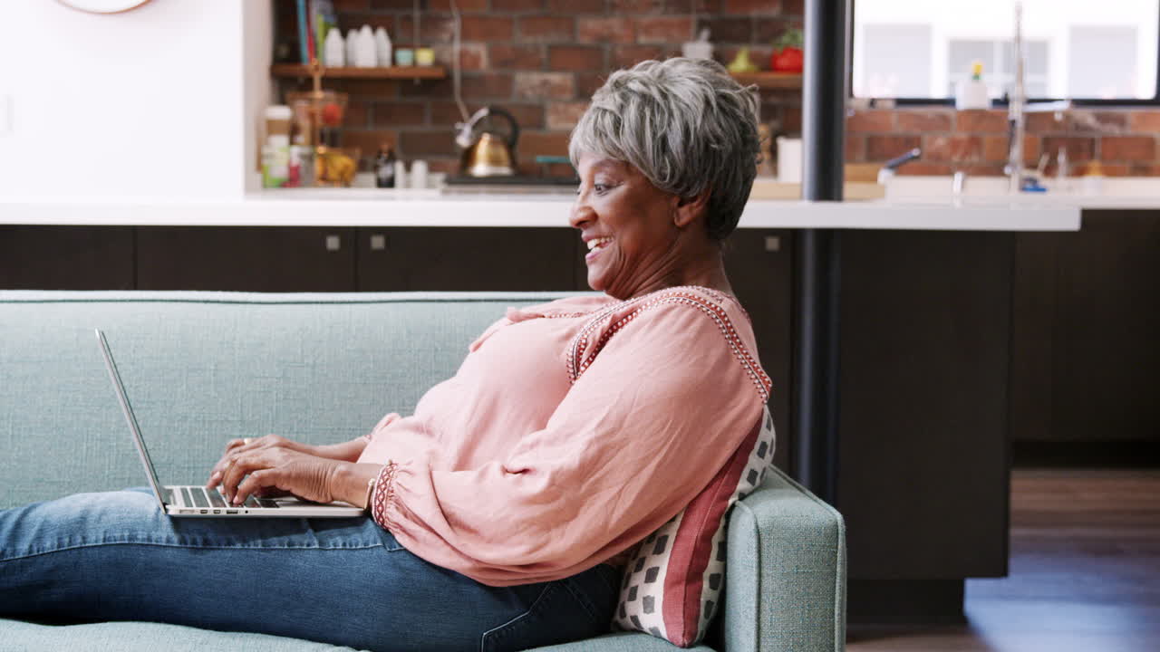 Senior Woman Relaxing On Sofa At Home Using Laptop To Shop Online