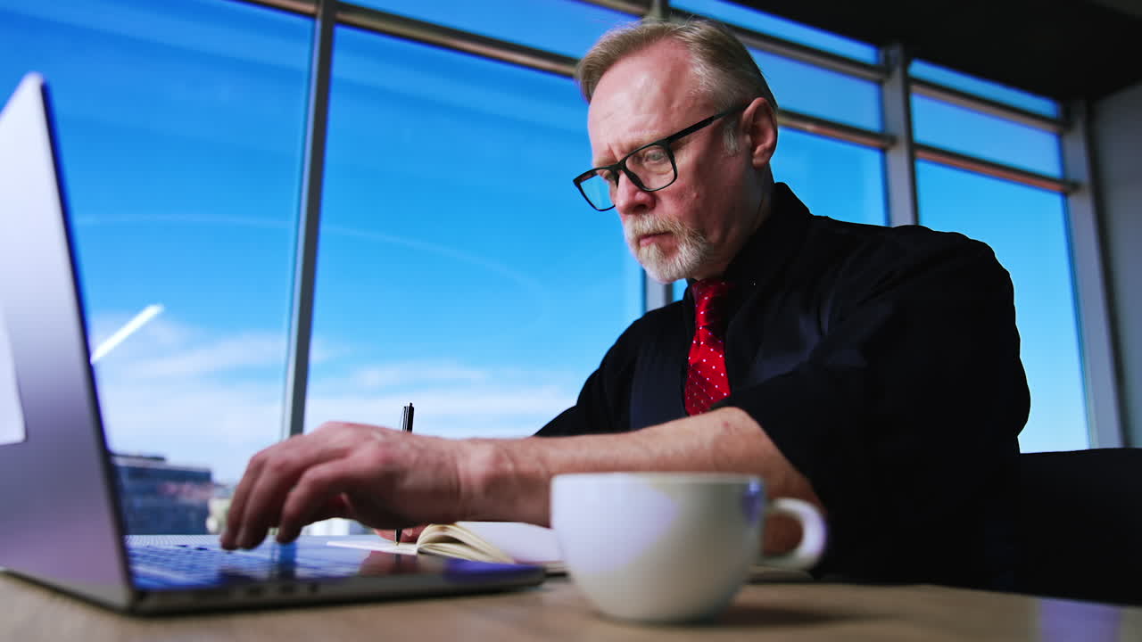 Hard-working senior businessman types on laptop and writes in his paper notebook. Man distracts on the coffee in white cup standing on the desk. Low angle view.