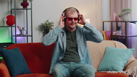 overjoyed young man in wireless headphones dancing, singing on cozy couch in living room at home