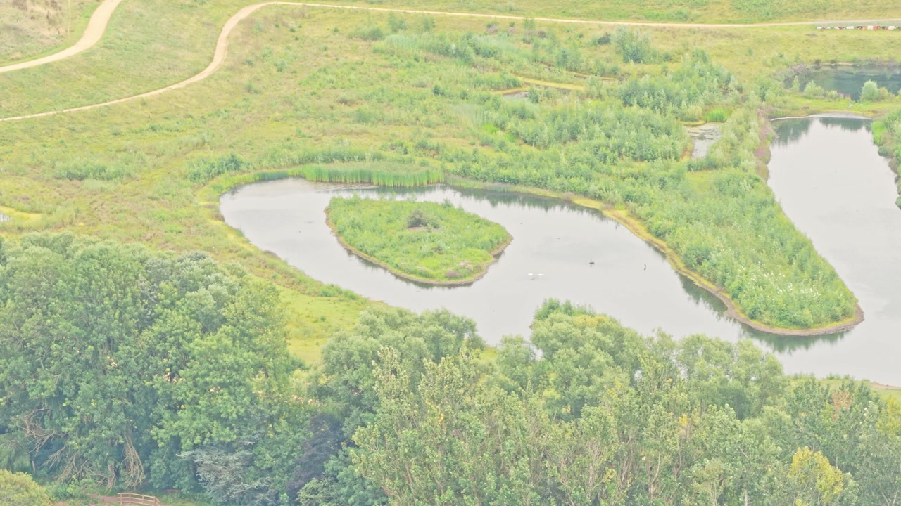 Curving pond with central vegetated island lies within the flood-defence wetlands of Kersal on the River Irwell’s old racecourse site in Salford, England, captured in elevated aerial view.