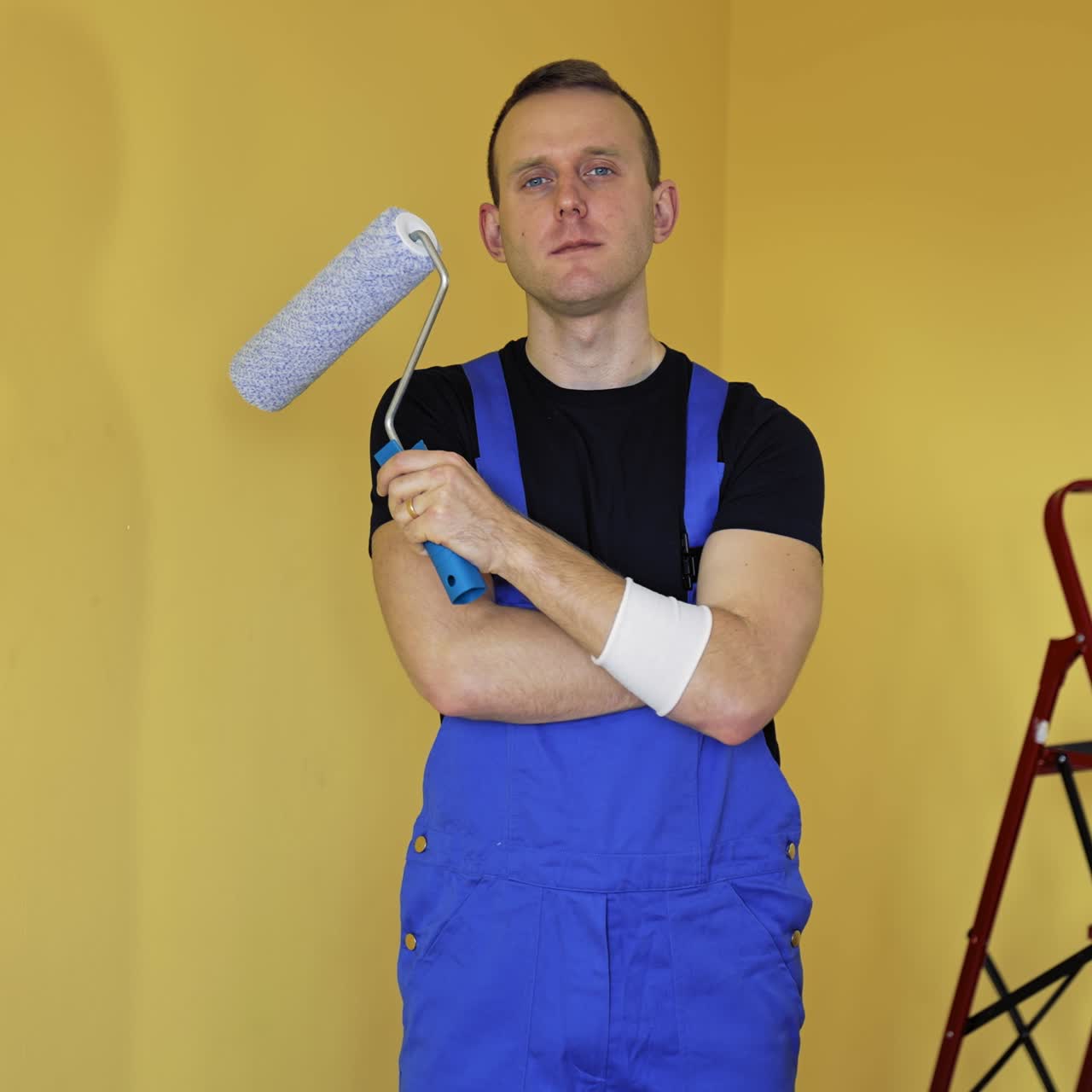 Portrait of a worker painter. Young man in blue overalls standing in the room and holding roller brush before doing a makeover in flat. Home improvement and renovation